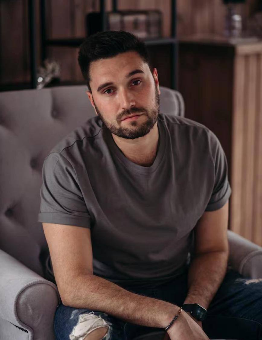 Man sitting on a couch wearing a gray t-shirt in a casual setting.